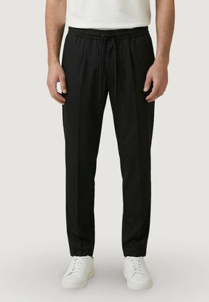 Uomo con pantaloni neri con coulisse e scarpe da ginnastica bianche, in piedi davanti a uno sfondo chiaro e semplice.