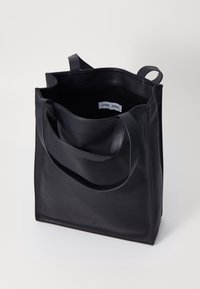 Sac fourre-tout en cuir noir avec une texture lisse, à ouverture sur le dessus et deux poignées. Présente un logo de marque en relief à l'avant. L'intérieur est non doublé.