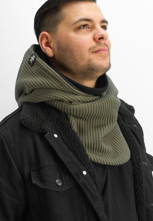 Knitted green scarf with ribbed texture and an adjustable size, worn over a black jacket with shearling collar. Features logo tag near the neckline.