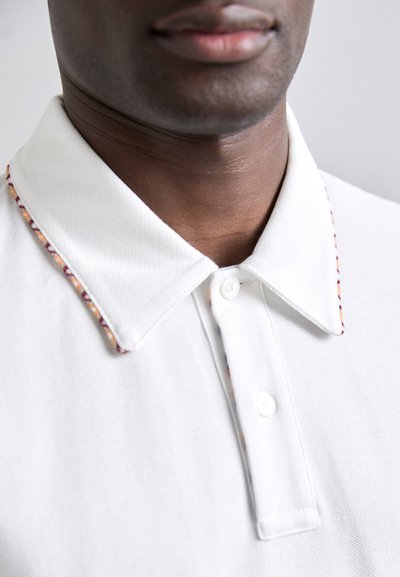 Κοντινή λήψη άσπρου πόλο μπλουζιού, με κλασικό γιακά και πολύχρωμες ραφές κατά μήκος των άκρων, ορατοί δύο κόμποι στο πανί.