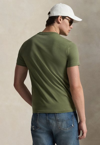 Πράσινο ελιάς κοντομάνικο μπλουζάκι από βαμβάκι, εφαρμοστή σχεδίαση, ομαλή υφή. Φορεμένο με μπλε τζιν με φθορές, εμφανίζεται από πίσω.