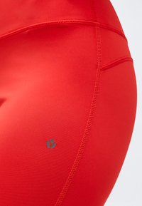 Gros plan sur des leggings de sport rouges mettant en évidence les détails des coutures et un petit logo réfléchissant sur le côté.