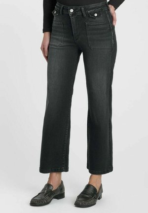 Donna che indossa jeans neri a vita alta, dritti, con tasche applicate frontali e mocassini neri, in piedi su sfondo bianco.