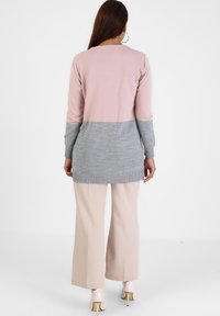 Maglietta a maniche lunghe in maglia con una sezione superiore rosa e una sezione inferiore grigia, con polsini a coste. Abbinata a pantaloni larghi beige chiaro.