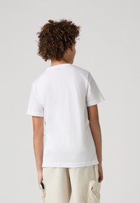 Camiseta blanca de algodón con mangas cortas, cuello redondo y espalda lisa. Llevada con pantalones cargo de un beis claro, que tienen bolsillos y un teléfono móvil.