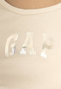 Débardeur côtelé beige clair avec lettrage "GAP" argent métallique au centre, doté d'un col rond et d'un tissu texturé.