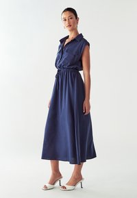 Abito camicia in lino blu navy con colletto, maniche corte arrotolate, vita stretta e gonna fluida lunga fino alla caviglia; presenta una tasca davanti.