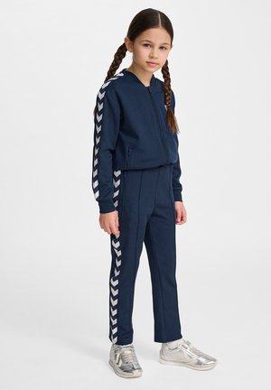 Mörkblå träningsoverall med vita chevronmönster på ärmar och sidor, bestående av en jacka med dragkedja och raka byxor. Silverfärgade sneakers.