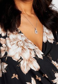 Tela negra con flores grandes en tono beige; presenta un profundo escote en V. Se ve un collar de cadena plateado con un colgante rectangular.