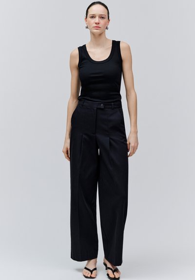 Vrouw staande tegen een grijze achtergrond, gekleed in een zwarte mouwloze top, zwarte broek met hoge taille en wijde pijpen, en zwarte sandalen, kijkend naar voren.