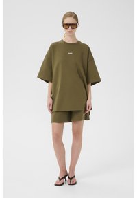 Olijfgroene oversized t-shirt met korte mouwen en logodetail, gecombineerd met bijpassende shorts. Draag het met donkere zonnebrillen en zwarte sandalen.