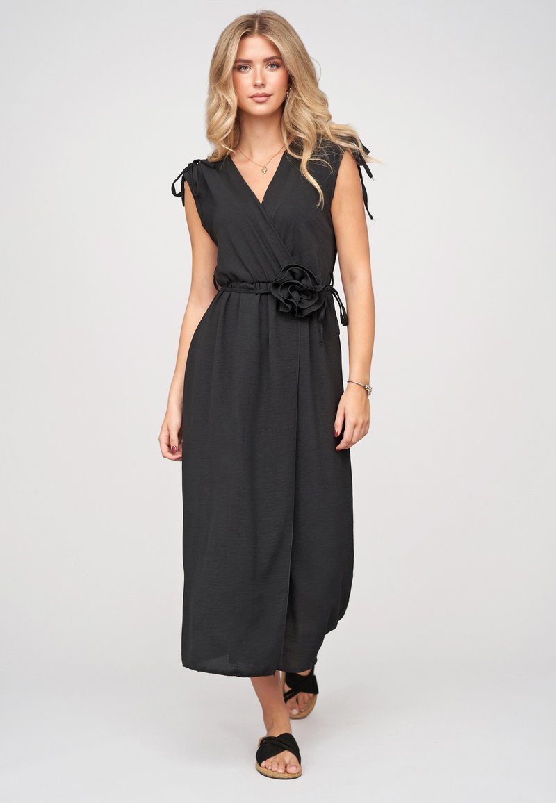 Femme blonde portant une robe portefeuille noire sans manches avec une ceinture fleur en tissu, marchant vers l'avant sur un fond clair uni.