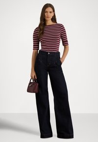 Mulher vestindo uma blusa às riscas bordeaux e branca com mangas até ao cotovelo, jeans escuros de cintura alta e perna larga, segurando uma pequena carteira bordeaux.