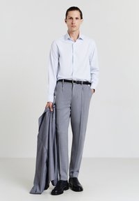 Hellblaues Hemd mit Knöpfen, graue Anzughose, schwarzer Gürtel und schwarze Schuhe. Hält einen passenden grauen Blazer. Schlichter Hintergrund.