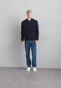 Jovem com cabelo loiro está de pé contra uma parede cinza, vestindo um cardigã preto abotoado, jeans azuis e tênis brancos.