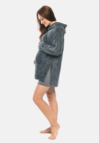 Flauschiger, dunkelgrauer Kapuzenmantel mit langen Ärmeln, Seitenschlitzen und einer Fronttasche. Hergestellt aus weichem, plüschigem Material. Modell trägt graue Shorts.