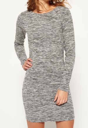 Kobieta ubrana w dopasowaną, długą, szarą melanżową sukienkę dzianinową, stojąca z jedną ręką na biodrze, a drugą luźno opuszczoną wzdłuż ciała.