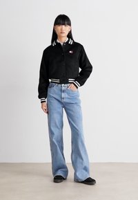 Sort cropped bomberjakke med hvide stribede manchetter og krave, parret med lyseblå brede bukser og sorte flade sko.