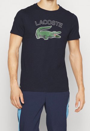 Mężczyzna ubrany w czarną koszulkę Lacoste z dużym zielonym logo krokodyla oraz granatowe spodnie z jasnoniebieskimi paskami po bokach.