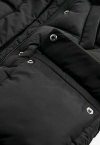 Gros plan d'une poche de veste matelassée noire avec des boutons-pression en métal et une fermeture éclair visible le long du bord.