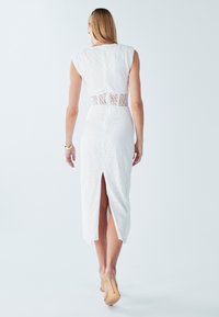 Robe en dentelle blanche avec manches courtes et insert en dentelle transparente à la taille. Le modèle comprend une fente au dos et une silhouette ajustée.