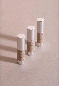 Tres envases cosméticos cilíndricos en plata y beige. Las etiquetas muestran el nombre del producto y el volumen (15 ml, 8 ml) en una fuente clara y elegante.