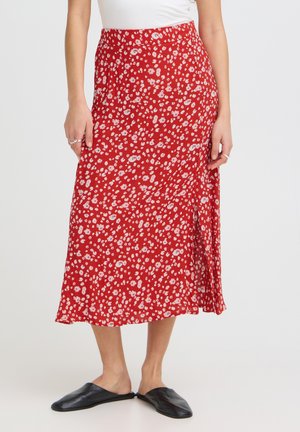 Femme portant une jupe midi rouge avec un petit motif floral blanc, un haut blanc, des bagues argentées et des chaussures plates noires à enfiler, debout sur un sol blanc.