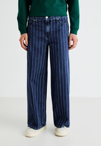 Blue denim jeans with white vertical stripes, wide-legged design, finished with a button closure at the waistband. Worn with white shoes.