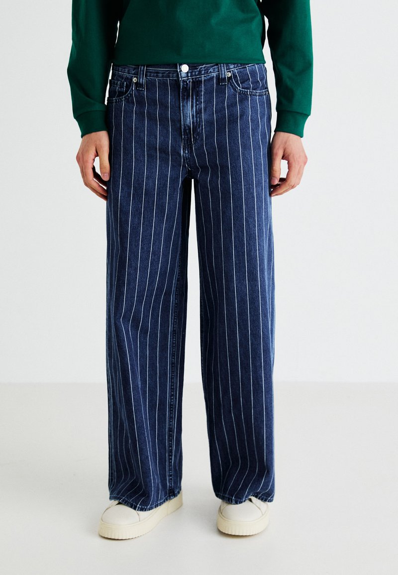 Blue denim jeans with white vertical stripes, wide-legged design, finished with a button closure at the waistband. Worn with white shoes.