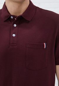Burgundia värvi polo särk tekstureeritud kudumisega, kahe nööbiga kaelas ja rinnataskuga, millel on triibuline aktsent punases, valges ja sinises.