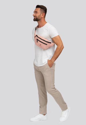 Roze heuptasje met zwarte ritsen, een gladde textuur en een logo, gedragen over de borst boven een wit shirt en beige broeken.