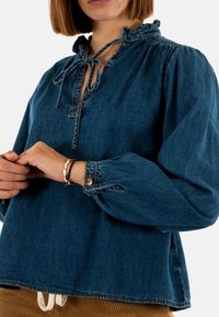 Blusa in denim blu con vestibilità ampia, scollatura arricciata, chiusura con fiocco e maniche a palloncino con dettagli di bottoni ai polsini.