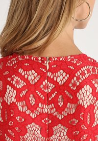 Robe rouge en dentelle présentant des motifs floraux ajourés, avec une fermeture éclair discrète au dos et une doublure beige contrastante.