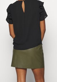 Schwarze Bluse mit kurzen, gerafften Ärmeln und einem Schlüsselloch-Detail hinten; kombiniert mit einem glatten Minirock aus olivegrünem Kunstleder.