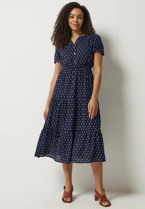Vrouw staand in navy midi-jurk met wit rechthoekig stippenpatroon, korte pofmouwen, geknoopte lijfje en bruine sandalen met hak.