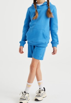 Haut à capuche en coton bleu avec poche kangourou, associé à un short bleu assorti. Le modèle porte des chaussettes blanches à motifs et des baskets avec des accents noirs.