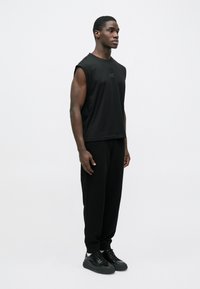 Fekete ujjatlan póló finom logóval, laza fekete nadrággal és fekete tornacipővel, mindez egyszerű fehér háttér előtt.