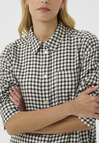 Une femme aux cheveux châtain clair porte une chemise à carreaux noir et blanc, les bras croisés, avec une expression neutre sur fond uni.