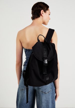 Personne portant un haut bustier et un jean avec un grand sac à dos noir muni d'une boucle frontale, vue de dos sur un fond uni.