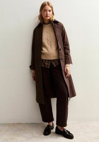 Ung kvinna iklädd brun trenchcoat, beige kabelstickad tröja, rutigt skjorta, mörkbruna byxor och svarta loafers, stående mot vit vägg.