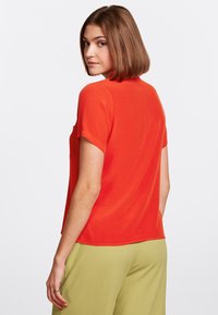 Top a maniche corte, di un vivace rosso, con una finitura texturizzata, caratterizzato da un collo rotondo e una vestibilità leggermente ampia, indossato con pantaloni verdi.