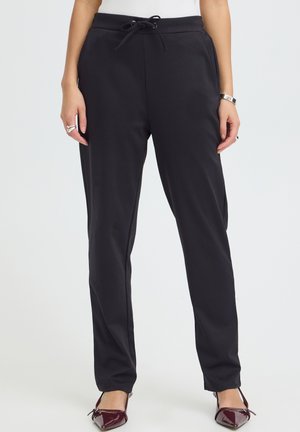 Personne portant un pantalon noir à cordon, un haut blanc, des bijoux en argent et des ballerines bordeaux, debout devant un fond clair et uni.