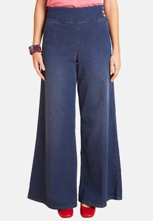 Frau trägt hoch taillierte, weit geschnittene dunkelblaue Jeans kombiniert mit einem pinken Top, buntem Armband und roten Schuhen mit quadratischer Spitze.