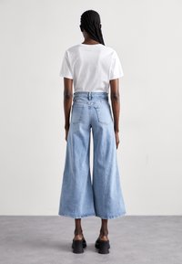 Pantalones vaqueros de tiro alto y pierna ancha de color azul claro, hechos de denim, con un dobladillo sin rematar. Combinados con una camiseta blanca sencilla, vistos desde atrás.