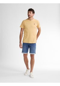 T-shirt di cotone giallo con maniche corte e un piccolo logo, abbinato a pantaloni corti in denim blu risvoltati all'orlo e sneakers bianche.