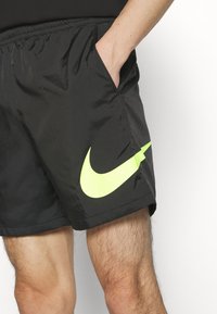 Hombre vistiendo pantalones cortos deportivos negros con un gran logo Nike en neón amarillo, con la mano en el bolsillo, de pie frente a un fondo blanco.