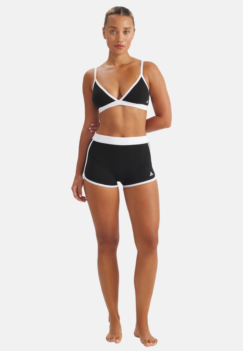 Femme debout pieds nus portant un soutien-gorge de sport noir et un short assorti avec une bordure blanche sur un fond blanc uni.