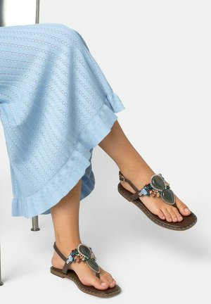 Piedi che indossano sandali marroni con grandi pietre verdi e perline colorate, abbinati a una gonna azzurro chiaro con trama.