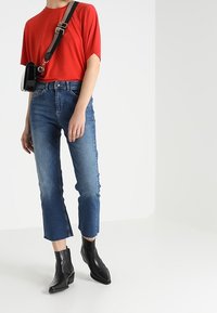 Top rosso a maniche corte, jeans blu a vita alta con orlo grezzo e stivali neri alla caviglia. Borsa a tracolla nera con tracolla marrone.