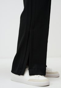 Czarne spodnie z bocznymi rozcięciami, wykonane z gładkiego materiału. Połączone z białymi trampkami o teksturowanej podeszwie i minimalistycznym designie.